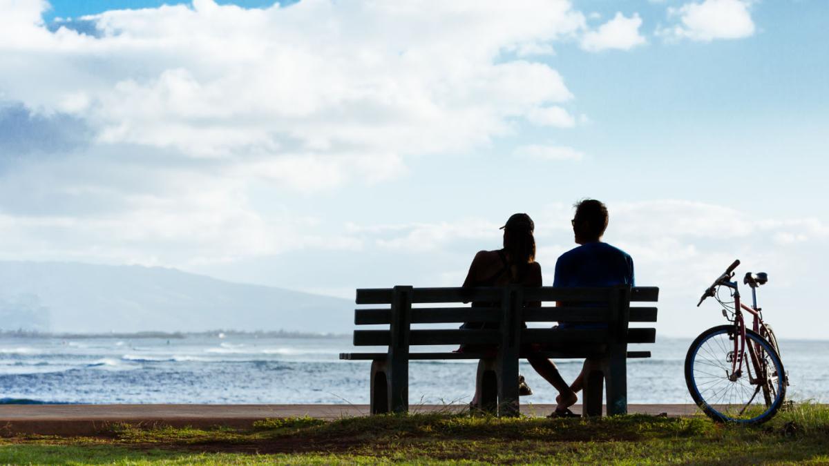 A couple sit on a bench along a coastline with their bicycles