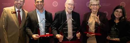 A distinguished college dean and four well-dressed award recipients stand together holding their awards at a formal event.